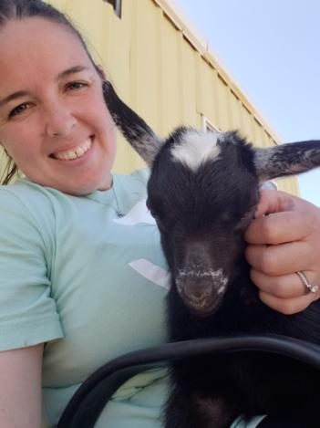A smiling woman holding a black and white goat kid outside the barn