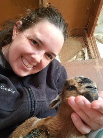 A smiling woman cradling a newborn brown goat kid inside the barn