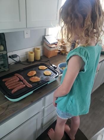A young girl standing on a stool cooking pancakes and sausage on a griddle
